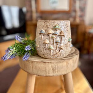 May include: A beige ceramic vase with a textured surface and raised mushroom designs in shades of tan, blue, and green. The vase is displayed on a small wooden stool, with a sprig of lavender in the foreground.