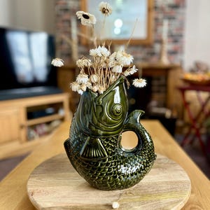 May include: A green ceramic fish-shaped vase with dried white flowers. The vase has a textured scale pattern and a handle. It sits on a wooden surface. The vase is a decorative item.