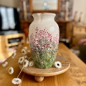 May include: A cream-colored ceramic vase with a floral design. The vase features a crackled finish and is adorned with red flowers and green foliage. The vase is displayed on a wooden pedestal.