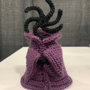 以下が含まれることがあります： 手作りのかぎ針編みのフィギュア。濃い紫色のマントと、上部に黒い渦巻き模様があります。このフィギュアは、目に見えるステッチで作成されており、白い表面に置かれ、糸の質感と詳細を示しています。