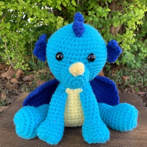 May include: A handmade crochet dragon plushie in shades of blue and yellow. The dragon has a blue head with dark blue accents, black eyes, and a yellow beak. The body is a mix of blue and yellow, with blue wings and feet. The plushie is sitting on a wooden surface.