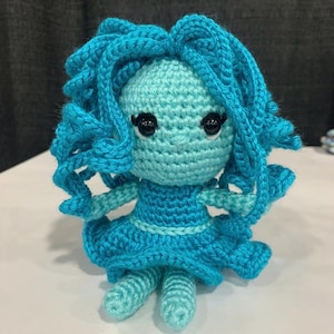 May include: A handmade crochet doll with a light blue body, turquoise hair, and a matching dress. The doll has black eyes and is posed on a white surface. The doll's hair is styled in curly ringlets.