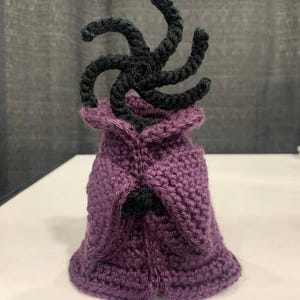 以下が含まれることがあります： 紫色のマントのようなベースと、黒い渦巻き状の触手を持つ手作りのかぎ針編み彫刻。彫刻はきついステッチで作られており、質感が生まれています。カラーパレットは紫と黒の色合いが中心です。
