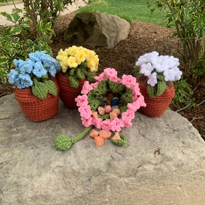 May include: A collection of handmade crochet flower pots in various colors, including blue, yellow, and white. Each pot features a detailed crochet flower arrangement and sits in a terracotta-colored pot. A pink flower basket with dice is also present.