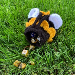 May include: A crocheted bee-shaped dice bag with black and yellow stripes and white wings. The bag is filled with colorful dice.