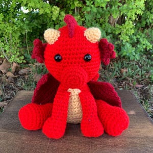 May include: A handmade red crochet dragon with black eyes, beige belly, and burgundy wings. The dragon has a small horn and beige ears. The dragon is sitting on a wooden surface with a green leafy background.