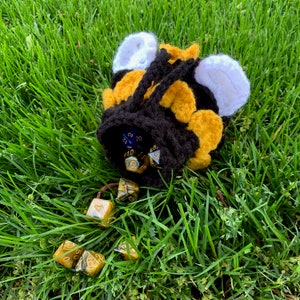 Puede incluir: Una bolsa de dados de crochet con forma de abeja con rayas negras y amarillas y alas blancas. La bolsa está llena de dados de seis caras de varios colores.