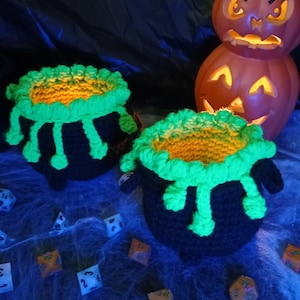 May include: Two black crocheted cauldrons with bright green textured rims featuring dripping accents. The interior of each cauldron is lined with orange yarn. A stack of three orange jack-o'-lanterns and several multi-sided dice are visible.