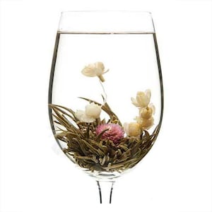 May include: A glass wine glass filled with water and a blooming tea flower. The flower is a light green color with white petals and a pink center. The tea flower is floating in the water.