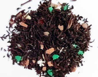 May include: A pile of loose leaf tea with visible pieces of cinnamon, and green and red tree-shaped sprinkles. The tea leaves are dark brown, and the sprinkles add a festive touch. The tea is set against a white background.