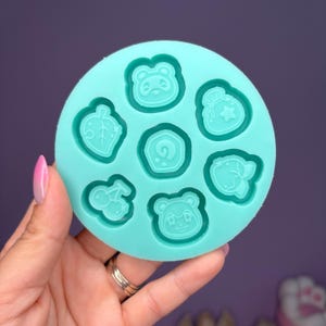 May include: A round, light teal silicone mold featuring seven different character designs. The designs include a leaf, a cherry, a bag of bells, a swirl, and three different animal faces. The mold is held in a hand with a pink painted nail.