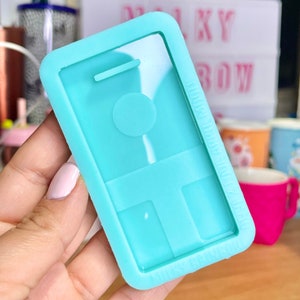 Puede incluir: Un molde de silicona azul claro con forma de teléfono inteligente con un círculo y dos rectángulos en el interior. El molde está diseñado para hacer manualidades de resina.