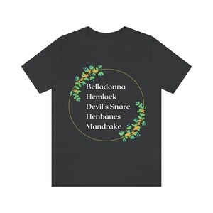 May include: Dark gray t-shirt featuring a gold circle with a floral design and the names "Belladonna, Hemlock, Devil's Snare, Henbanes, Mandrake" in white text. The shirt is a crew neck.