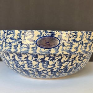 May include: A round ceramic bowl with a blue and white sponge-painted pattern. An oval blue sticker on the side reads: Hand Turned in the Ancient Tradition by Skilled Craftsman at Marshall Pottery. The bowl has a wide, flat rim and a deep basin.