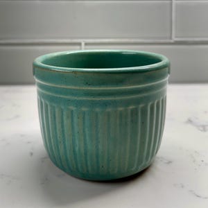 May include: A teal ceramic cup featuring a ribbed, vertical fluted texture along the exterior walls. The vessel has a smooth, rounded rim and a consistent, speckled glaze finish that covers the entire outer surface of the cylindrical container.