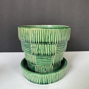 May include: A green ceramic plant pot with a matching saucer, featuring a textured basket-weave pattern with vertical and horizontal embossed lines. The pot has a tapered, tiered silhouette with a glossy finish and a wide, open circular rim.