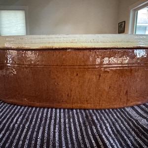 May include: An oval-shaped ceramic baking dish with a glossy brown glazed body and a contrasting cream-colored glazed rim. The vessel features two small, integrated side handles and a smooth, uniform exterior finish with subtle horizontal detailing.