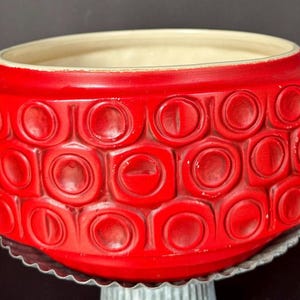 May include: A vibrant red ceramic planter with a repeating pattern of circles and squares. The planter has a wide, open top with a cream-colored interior. It sits on a decorative metal base.
