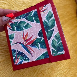 Pu&ograve; includere: Una scatola rossa con un motivo floreale rosa e verde. La scatola ha una patta con un'etichetta bianca. Il motivo floreale presenta foglie tropicali e fiori arancioni e gialli.