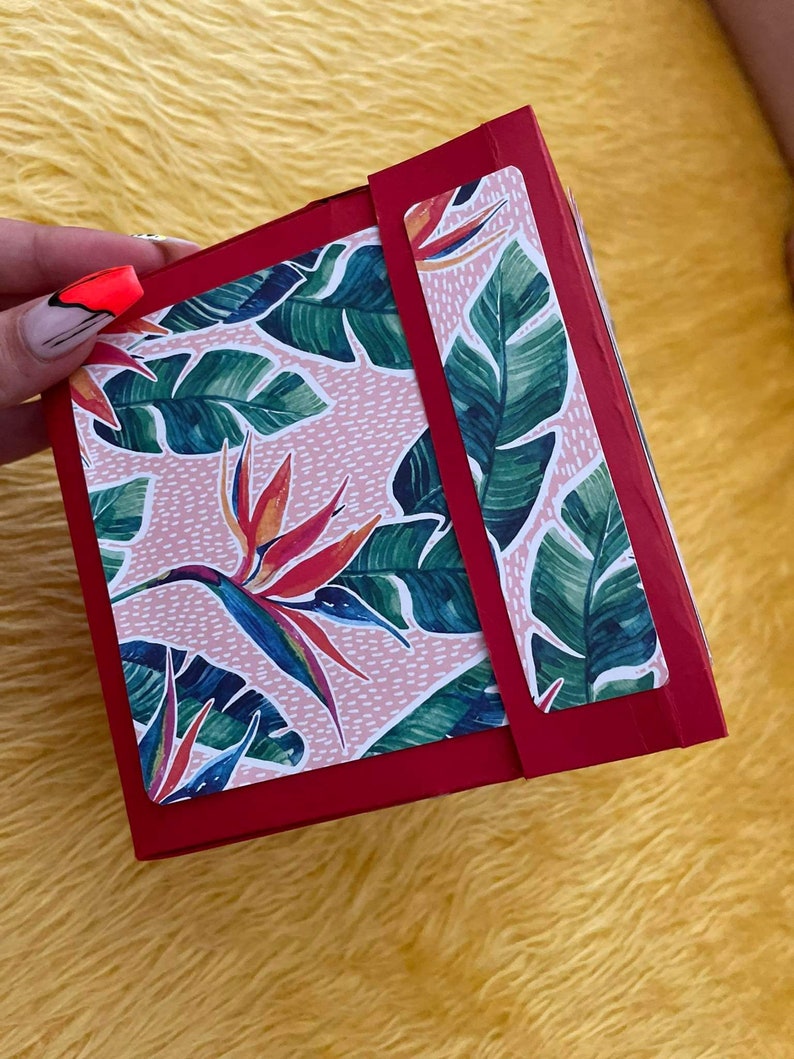 Pu&ograve; includere: Una scatola rossa con un motivo floreale rosa e verde. La scatola ha una patta con un'etichetta bianca. Il motivo floreale presenta foglie tropicali e fiori arancioni e gialli.