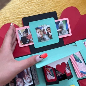 Pu&ograve; includere: Una scatola esplosiva blu turchese con accenti neri e rossi. La scatola ha pi&ugrave; scomparti con foto di due donne. La scatola ha uno sportello a forma di cuore nella parte superiore.