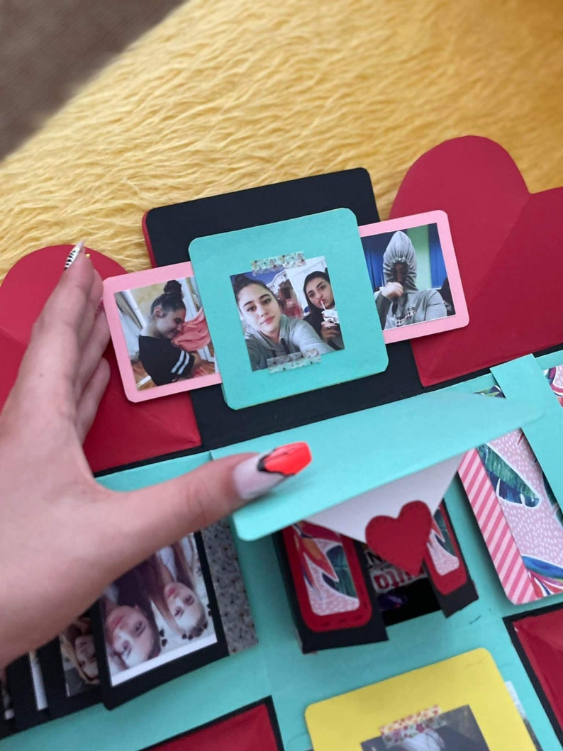 Pu&ograve; includere: Una scatola esplosiva blu turchese con accenti neri e rossi. La scatola ha pi&ugrave; scomparti con foto di due donne. La scatola ha uno sportello a forma di cuore nella parte superiore.