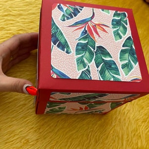 Pu&ograve; includere: Una scatola rossa con un motivo a foglie tropicali rosa e verdi sulla parte superiore e sui lati. La scatola &egrave; chiusa.