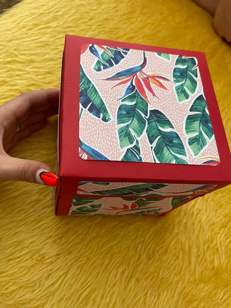 Pu&ograve; includere: Una scatola rossa con un motivo a foglie tropicali rosa e verdi sulla parte superiore e sui lati. La scatola &egrave; chiusa.