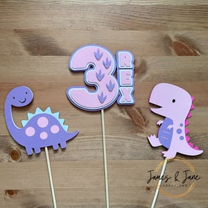 May include: Three pink and purple dinosaur cake toppers. The number three is pink with purple dinosaur footprints. The word "REX" is in purple letters. There are two purple dinosaurs, one with pink spots and one with pink stripes.