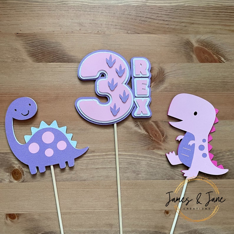 May include: Three pink and purple dinosaur cake toppers. The number three is pink with purple dinosaur footprints. The word "REX" is in purple letters. There are two purple dinosaurs, one with pink spots and one with pink stripes.
