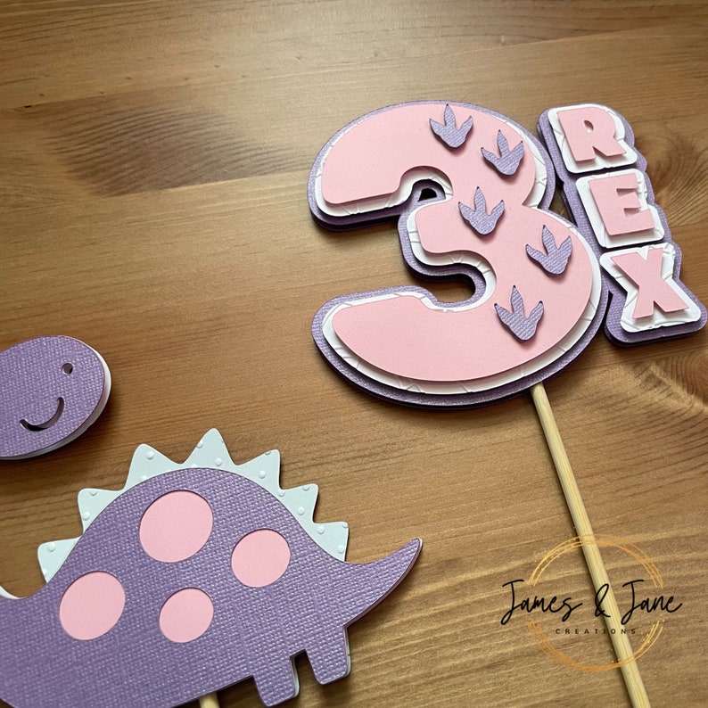 May include: Purple and pink dinosaur cake topper with the number 3 and the word "REX" on it. The number 3 has pink dinosaur footprints on it.