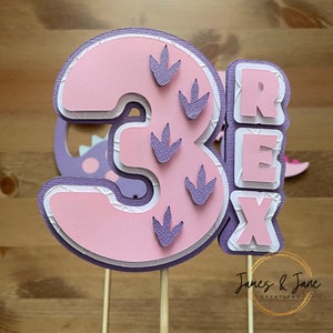 May include: Pink and purple layered paper cake topper with the number 3 and the word "REX" in white letters. The number 3 has purple dinosaur footprints on it.