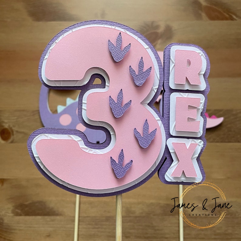 May include: Pink and purple layered paper cake topper with the number 3 and the word "REX" in white letters. The number 3 has purple dinosaur footprints on it.