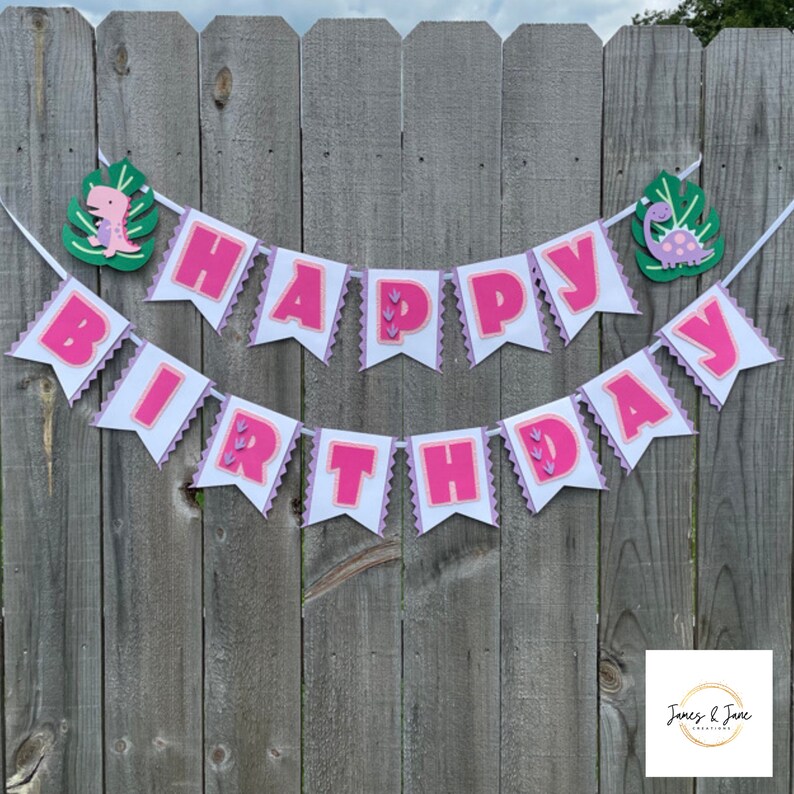 May include: Pink and white banner with the words "Happy Birthday" in pink letters. The banner is decorated with green leaves and pink dinosaurs.