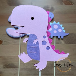 May include: Purple dinosaur cake topper with pink accents and a textured surface. The dinosaur is standing on a stick and has a small black eye. The topper is part of a set of three dinosaurs.