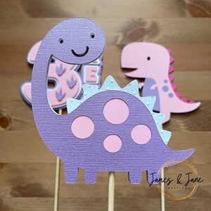 May include: Purple dinosaur cake topper with pink spots and a smiling face. The dinosaur has a light blue frill around its neck. The topper is on a wooden stick.  "James & Jane Creations" is printed on the bottom of the stick.