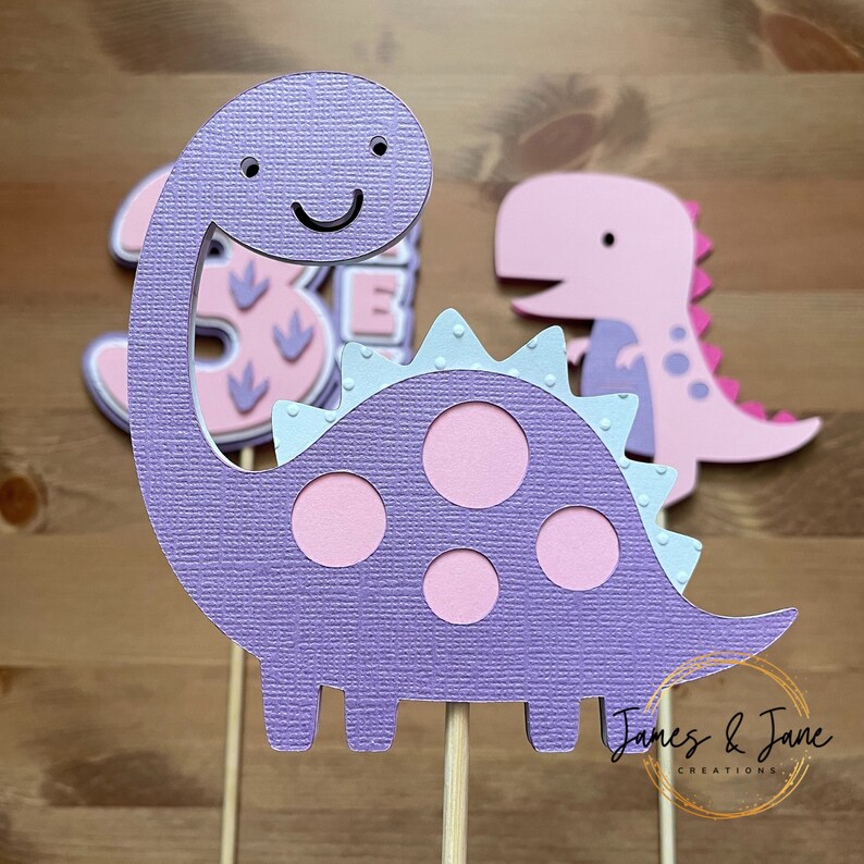 May include: Purple dinosaur cake topper with pink spots and a smiling face. The dinosaur has a light blue frill around its neck. The topper is on a wooden stick.  "James & Jane Creations" is printed on the bottom of the stick.