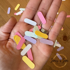 Peut inclure: Une collection de confettis rectangulaires colorés et à motifs dans différentes couleurs pastel, notamment rose, jaune, bleu et violet. Les confettis sont éparpillés sur une main.