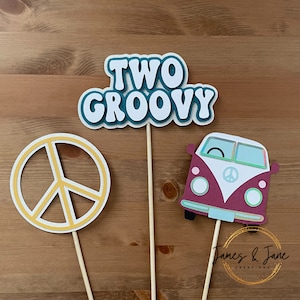 Può includere: Tre sagome di carta su bastoncini di legno. La prima sagoma è un cartello blu-verde e bianco con la scritta "TWO GROOVY". La seconda sagoma è un segno di pace giallo. La terza sagoma è un furgone Volkswagen color borgogna e bianco con un segno di pace sul davanti.