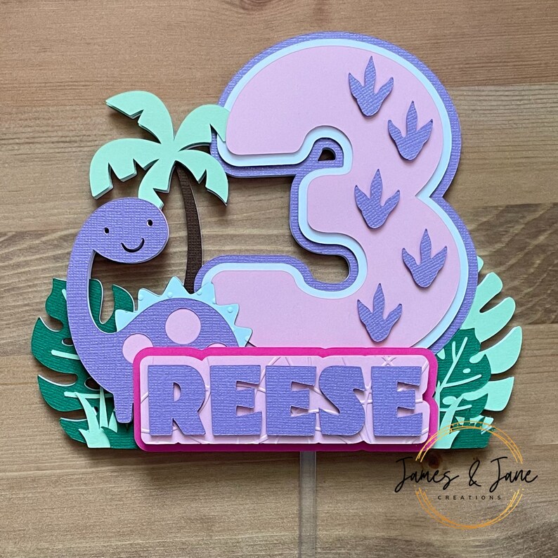 May include: Purple dinosaur cake topper with a pink number 3, green leaves, and the name "REESE" in purple letters.