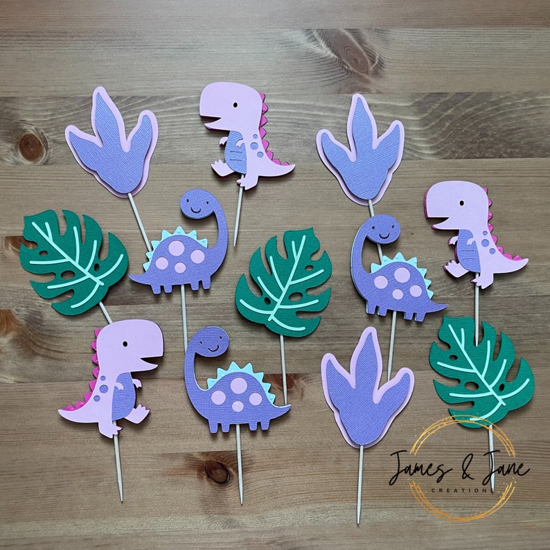 May include: Purple and pink dinosaur cupcake toppers with green leaves and purple footprints. The toppers are made of cardstock and have a whimsical design.