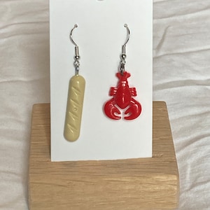 Può includere: Un paio di orecchini pendenti con un ciondolo a forma di aragosta rossa e una baguette bianca. Gli orecchini sono esposti su un supporto in legno.