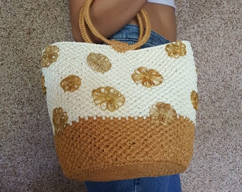 May include: A woven tote bag with a cream-colored top section adorned with tan flower appliqués and a tan bottom section. The bag has a circular handle and is held by a person.
