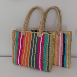 May include: Two rectangular crochet tote bags with sturdy beige handles. The front bag features vertical stripes in bright orange, hot pink, white, teal, and dark green. The back bag displays vertical stripes in yellow, pink, white, and dark blue.