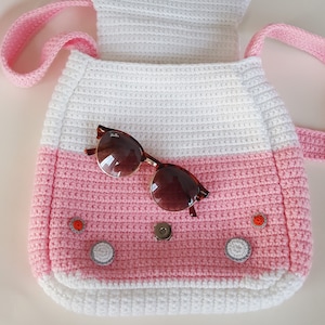 May include: A handmade crochet bag in white and pink, with a flap and a shoulder strap. The bag features decorative buttons and a magnetic closure. A pair of tortoiseshell sunglasses rests on the bag.