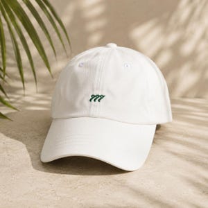 May include: White baseball cap with a curved brim. The cap features a small, embroidered green "777" design on the front. The hat is made of a soft material and has a classic, casual style. The background includes palm leaves and a sandy surface.