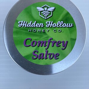 May include: A round, silver-colored tin with a green label featuring the text "Hidden Hollow Honey Co." and "Comfrey Salve" in purple. The label also includes a bee logo.