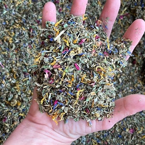 May include: A handful of dried herbs and flower petals, including blue, yellow, and pink, are held in an open hand. The mix appears to be a blend of various dried plants, with a focus on colorful floral elements. The background is filled with a similar mix.