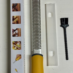 May include: A stainless steel zester grater with a yellow handle, protective cover, and cleaning brush. The grater is designed for fine grating and zesting. The packaging shows images of the grater in use.