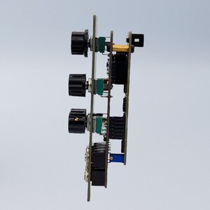 Pu&ograve; includere: Una vista laterale ravvicinata di una scheda a circuito stampato con tre manopole nere e un filo blu. La scheda ha uno sfondo verde ed &egrave; montata su una superficie bianca.
